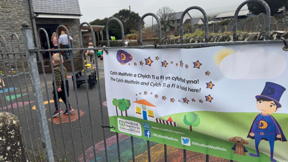 Llun o faner Mudiad Meithrin yn hongian ar gat y tu allan i gylch meithrin gyda plant yn chwarae tu allan. 