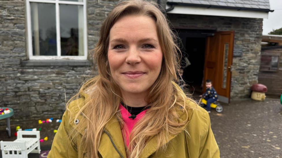 Dynes mewn cot felen gyda gwallt hir melyn yn gwenu ar y camera gyda tegannau a plant yn chwarae y tu ol iddi. 