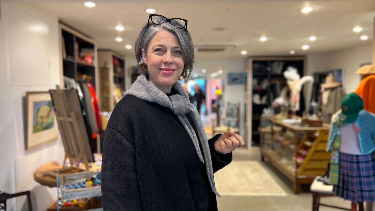 Bella Farrell stands inside a charity shop, which is warmly lit, with shelves and racks displaying a mix of clothing, accessories, and decorative items. She is wearing a dark coat with a light grey scarf tucked inside it. She has shorter grey hair and a pair of glasses resting on top of their head.