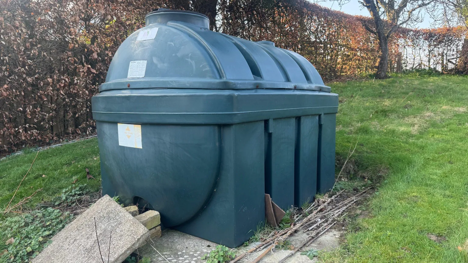 A large heating oil tank is in the centre of the image. It is surrounded by a copper beech tree hedge and a lawn 