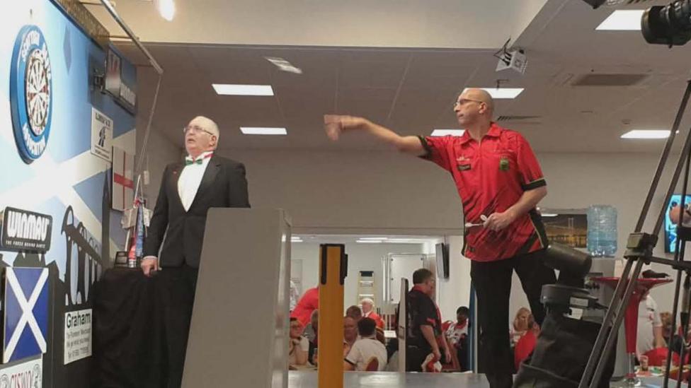 Clive Langford standing at the oche throwing a dart from right to left of the image
