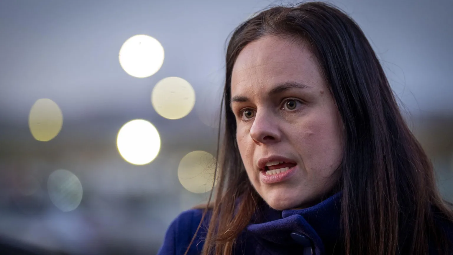  Kate Forbes has dark hair to her shoulders and is wearing a blue jacket. She is standing outdoors and there are blurred lights in the background.
