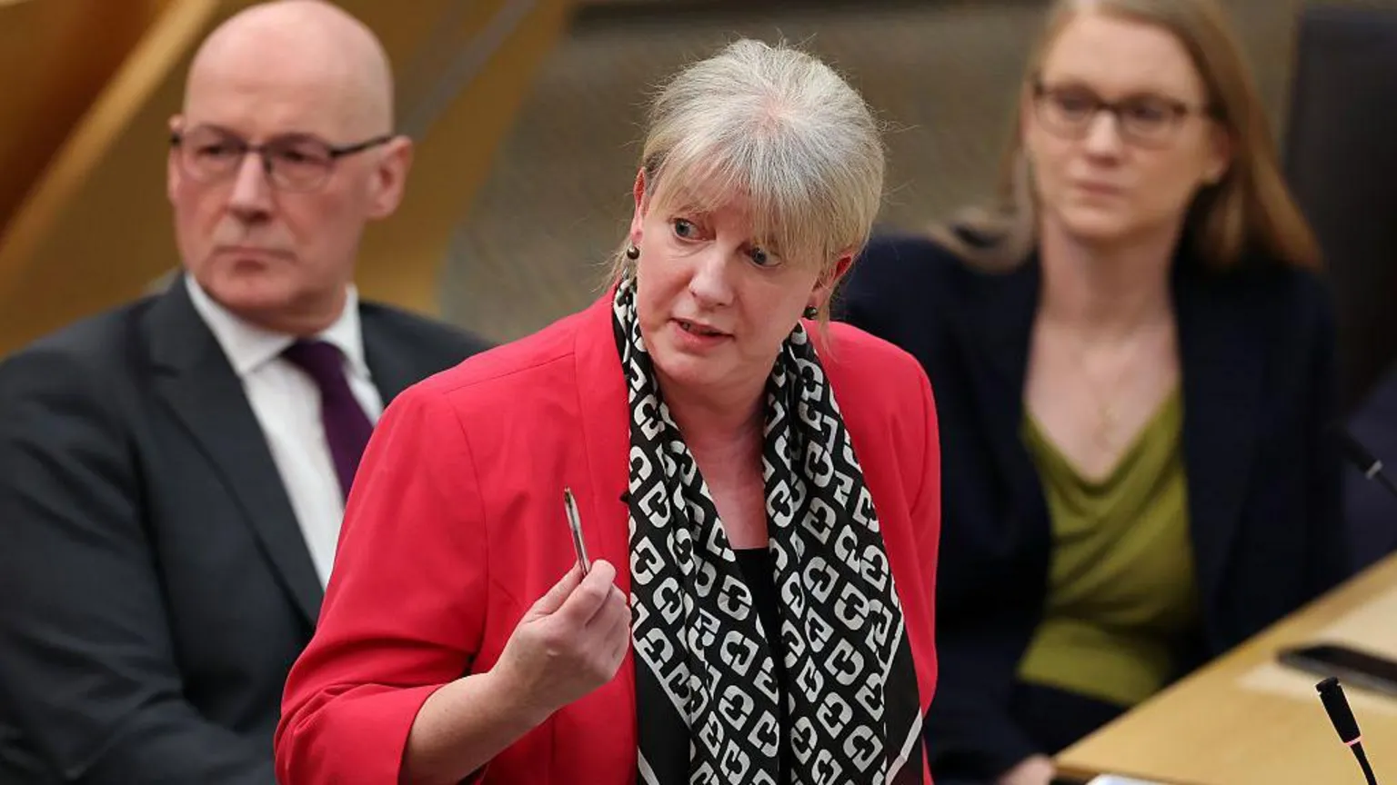  Scottish Finance Secretary Shona Robison - a middle-aged woman with blonde hair pulled back into a ponytail, wearing a red blazer with a black and white scarf - brandishes a pen while answering a question at Holyrood. First minister John Swinney and fellow minister Shirley-Anne Somerville look on in the background, slightly out of focus