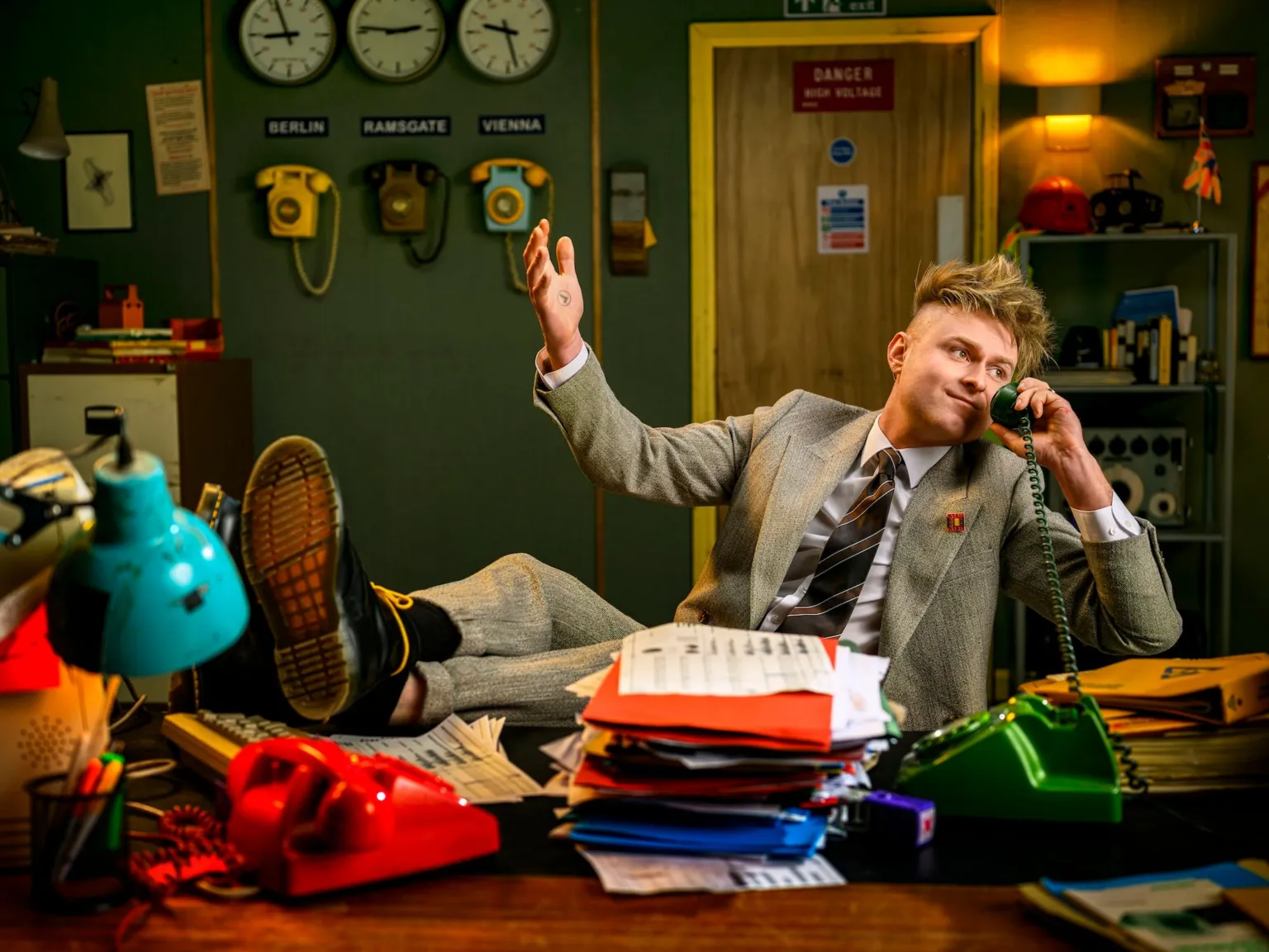 Look Mum No Computer holds a vintage telephone receiver to his ear, with his feet propped up on a desk, in a scene from the music video for Eins, Zwei, Drei