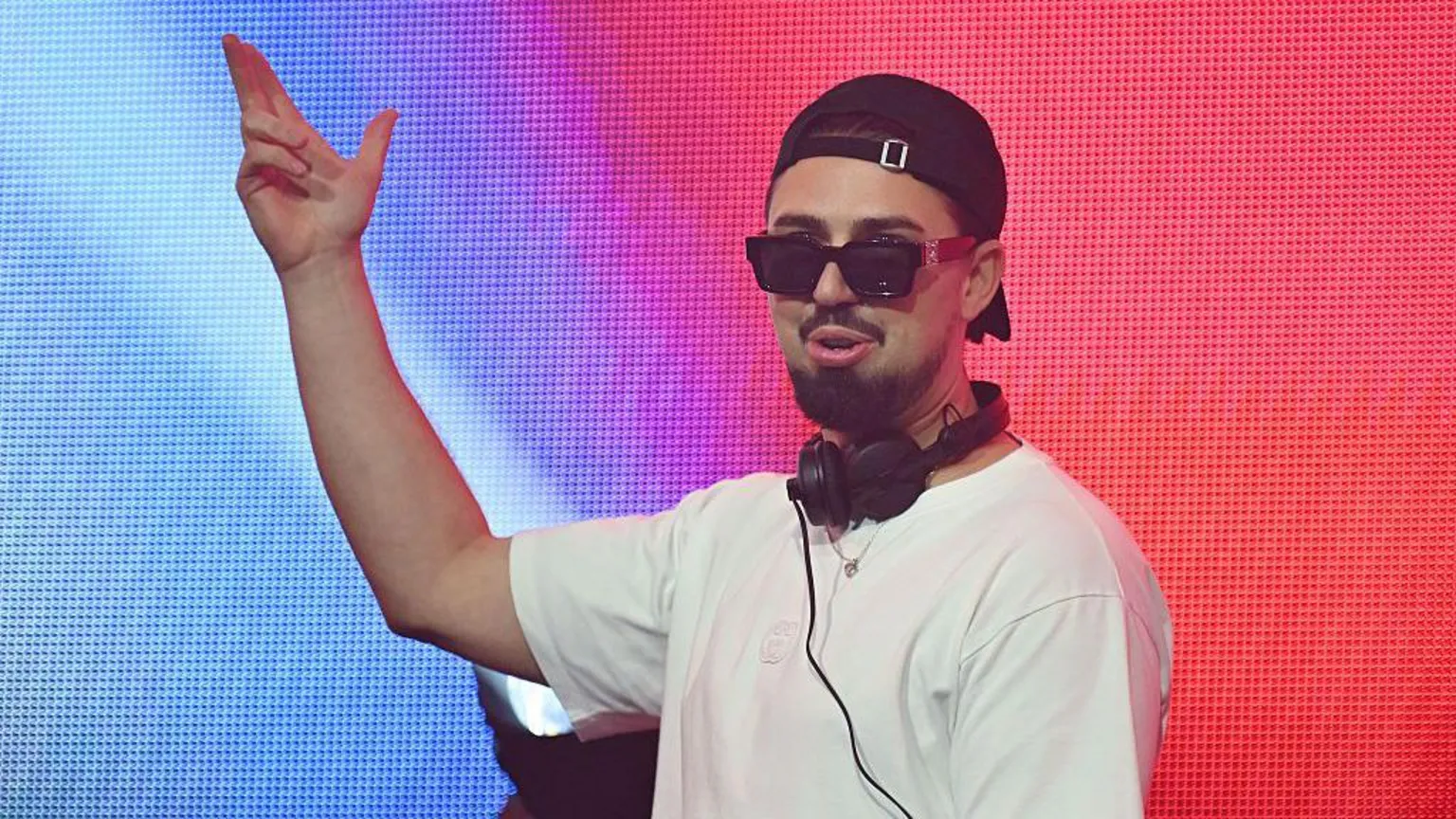  Sonny Fodera is wearing a white T-shirt and black cap with black headphones around his neck. 