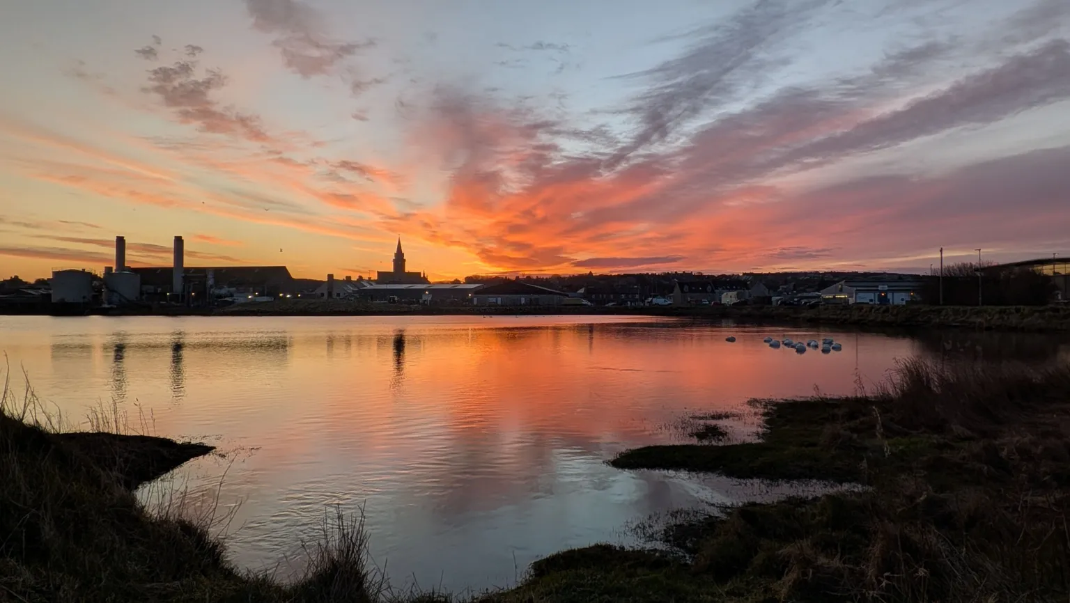 Sophie Blanchard A vivid sunset casts orange and pink reflections across a calm body of water, with a town skyline— including tall chimneys and a central tower— silhouetted against the sky.