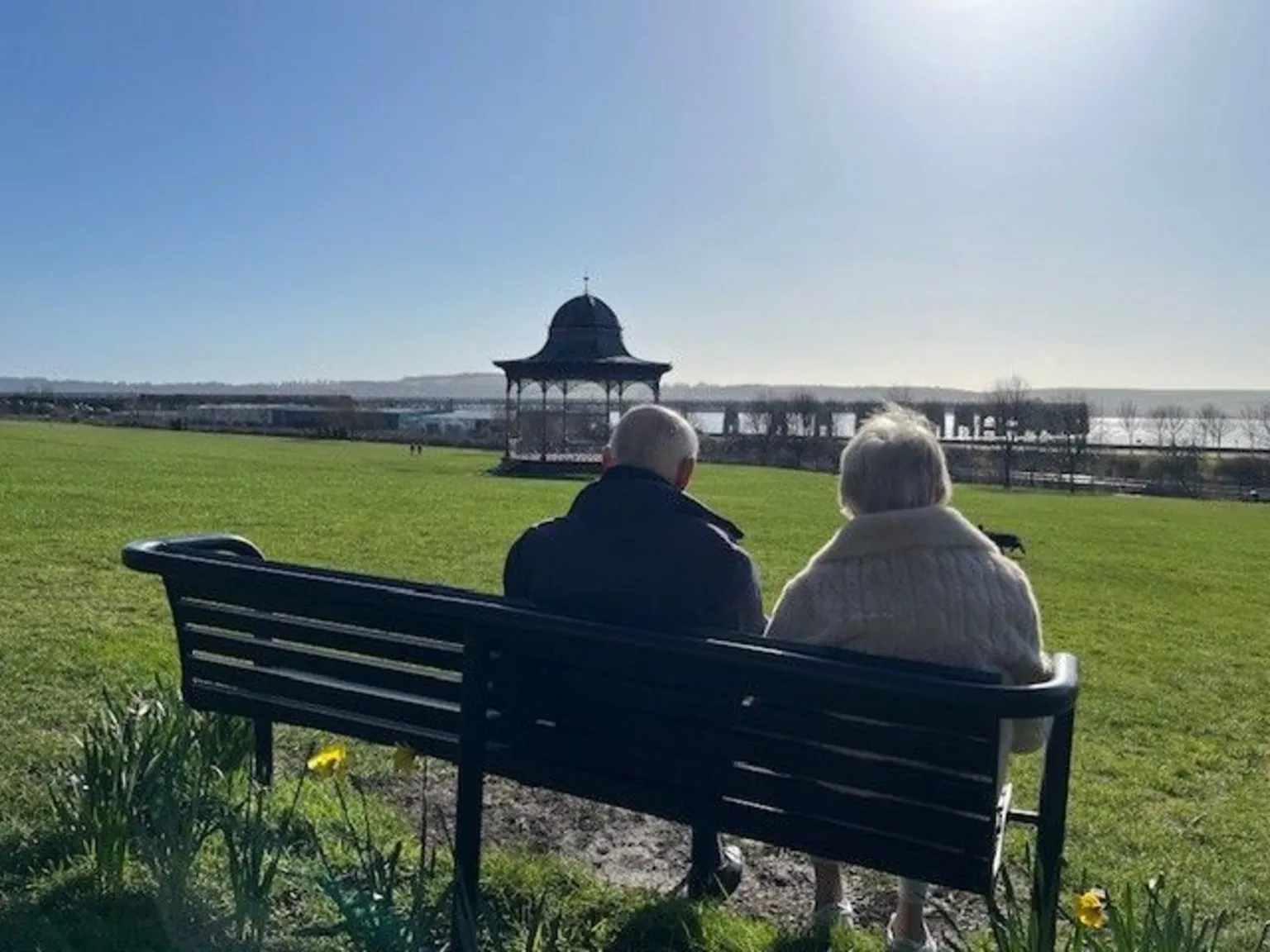 Elaine Morrison A wide grassy park on a sunny day, with two people sitting on a black bench facing away from the camera. In the distance stands an ornate bandstand and a large body of water. Daffodils bloom near the bench, and the sky is clear and bright.