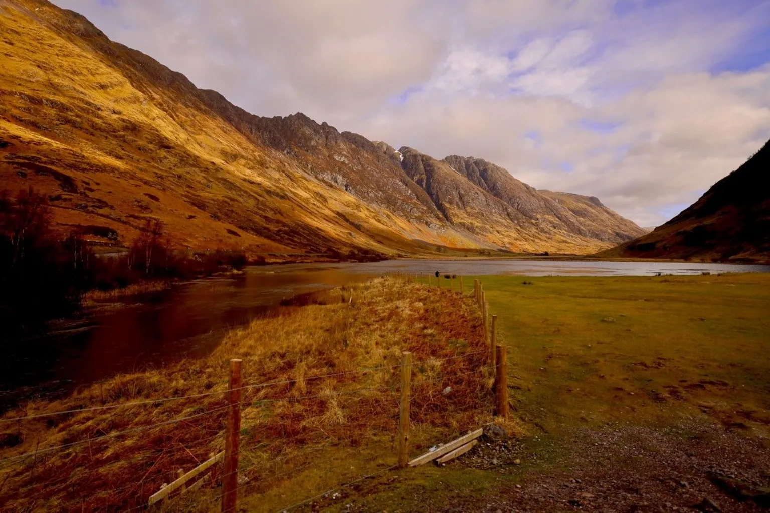 Graham Christie A broad valley surrounded by steep, golden‑brown mountains. A river runs along the left side, and grassy land stretches to the right. The sky is partly cloudy with sunlight casting warm tones across the scene.