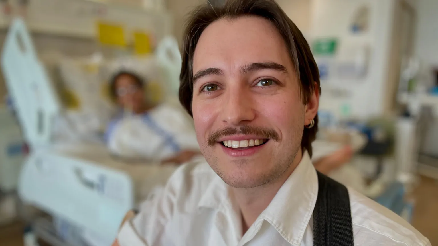 Mark Norman BBC Music therapist Joe Jezard smiles at the camera sitting in a hospital ward with one of his therapy patients in the background 