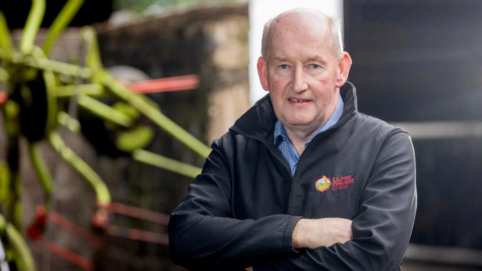 UFU Glenn Cuddy is bald and has his arms folded. He's wearing a black jacket that says Ulster Farmers' Union in red lettering on the left breast. Under the jacket he is wearing a blue checked shirt.