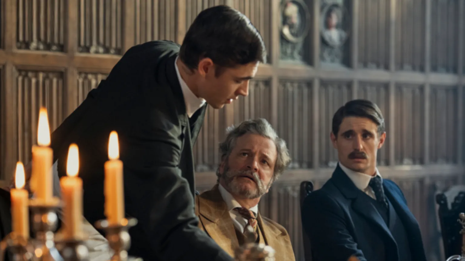 Daniel Smith/ Amazon MGM Studios Colin Firth has white hair and beard. He wears a beige three peice suit. He is sat down, and looking up at Holmes. Another character in a blue suit is sat next to Firth on the right. Four lit candlesticks are on the left.