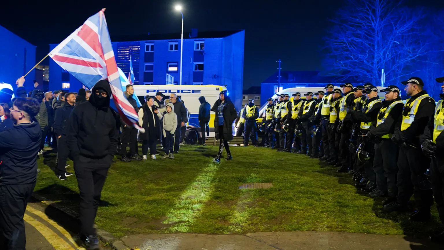 Police officers form a line in front of a group of protesters with police vans in the background
