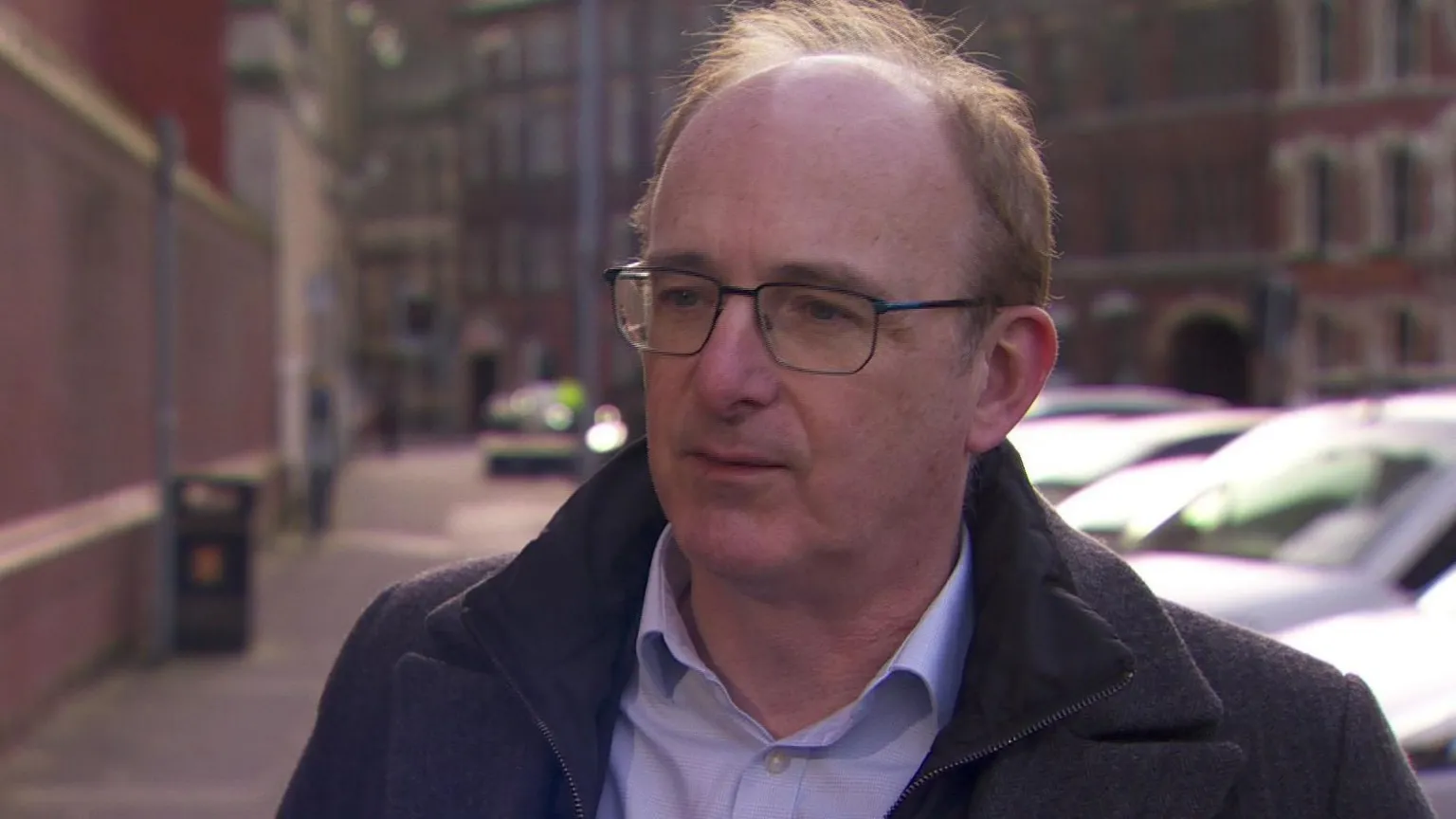 David Rooney has short brown hair and black-framed glasses. He is wearing a heavy coat over a shirt that is open at the neck