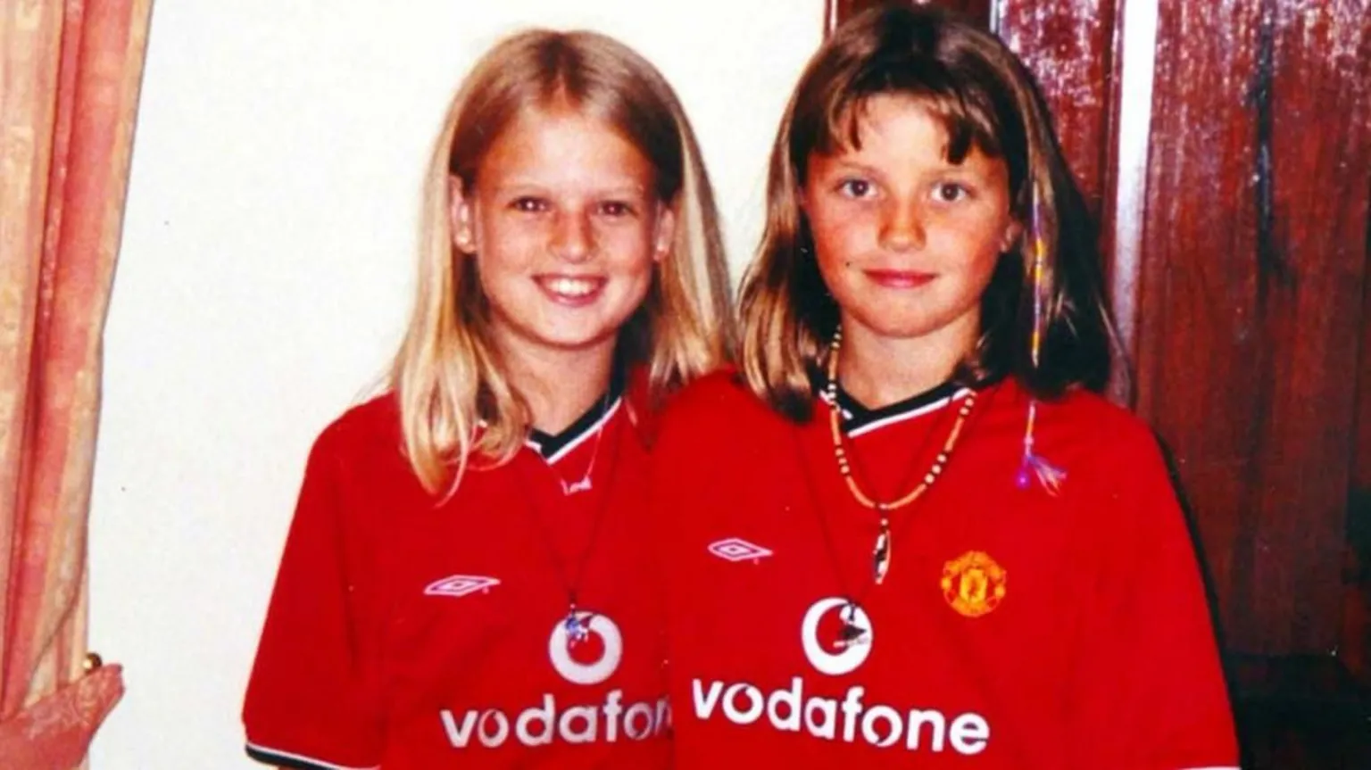  Two young girls dressed in matching red Manchester United shirts, smiling at the camera. 