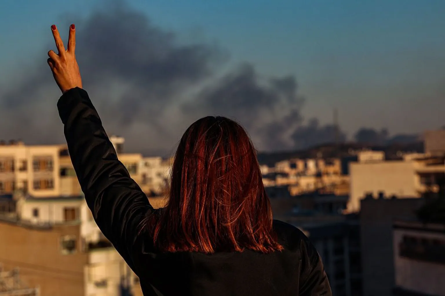  A woman holds a two-finger peace sign, while she stands on a rooftop as smoke rises in the distance over Tehran on 3 March 2026.