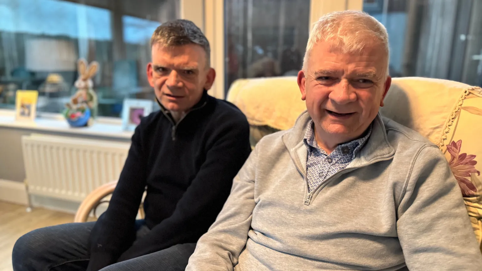A man with greying hair and a black jumper sits on a sofa with his hands between his legs. On his right a grey haired man with a grey half zip sits beside him