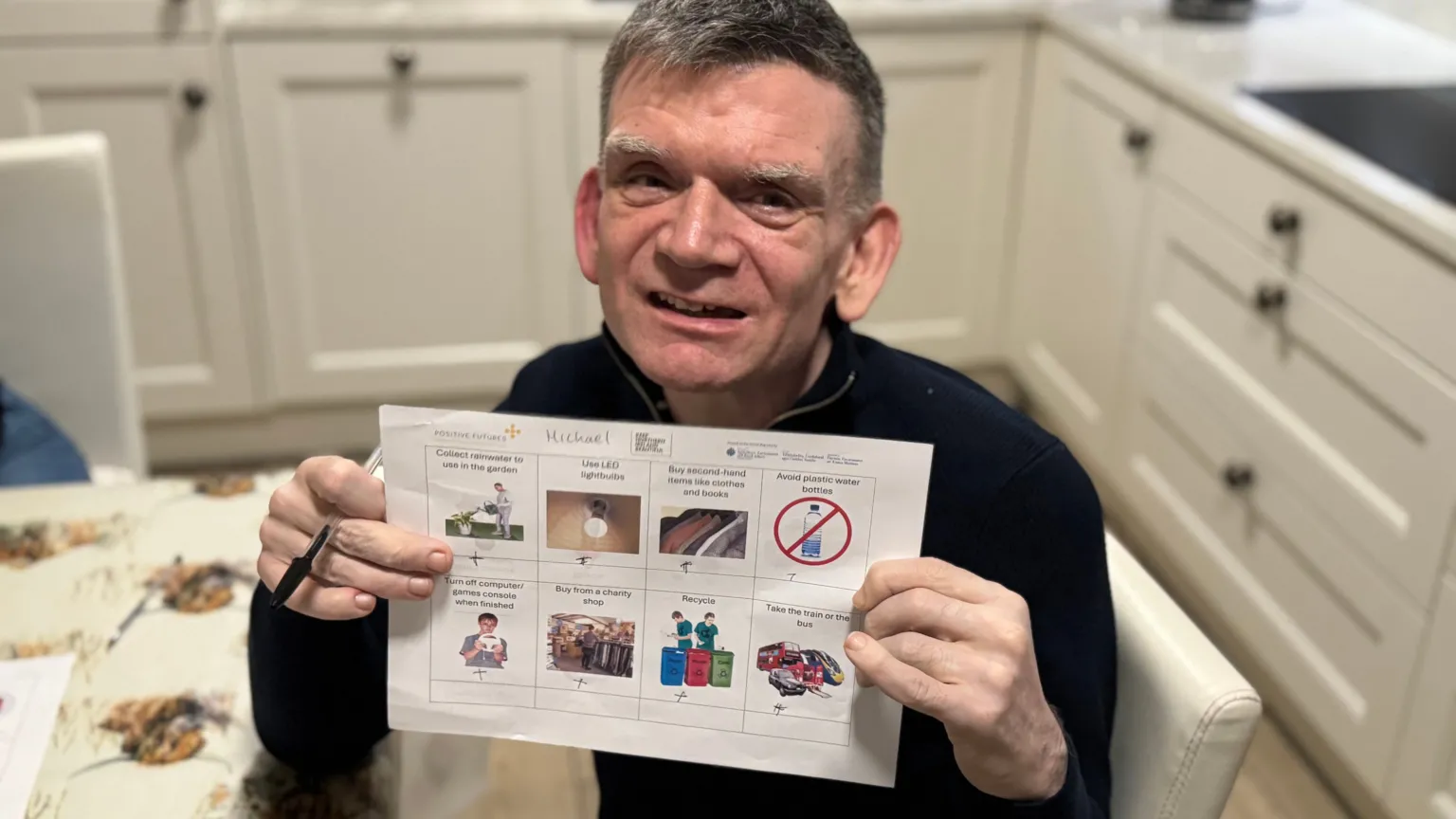 A man with greying hair and a black jumper on holds up a sheet with words and pictures on it. He also holds a pen in his hand. In the background you can see he is sitting in a kitchen 