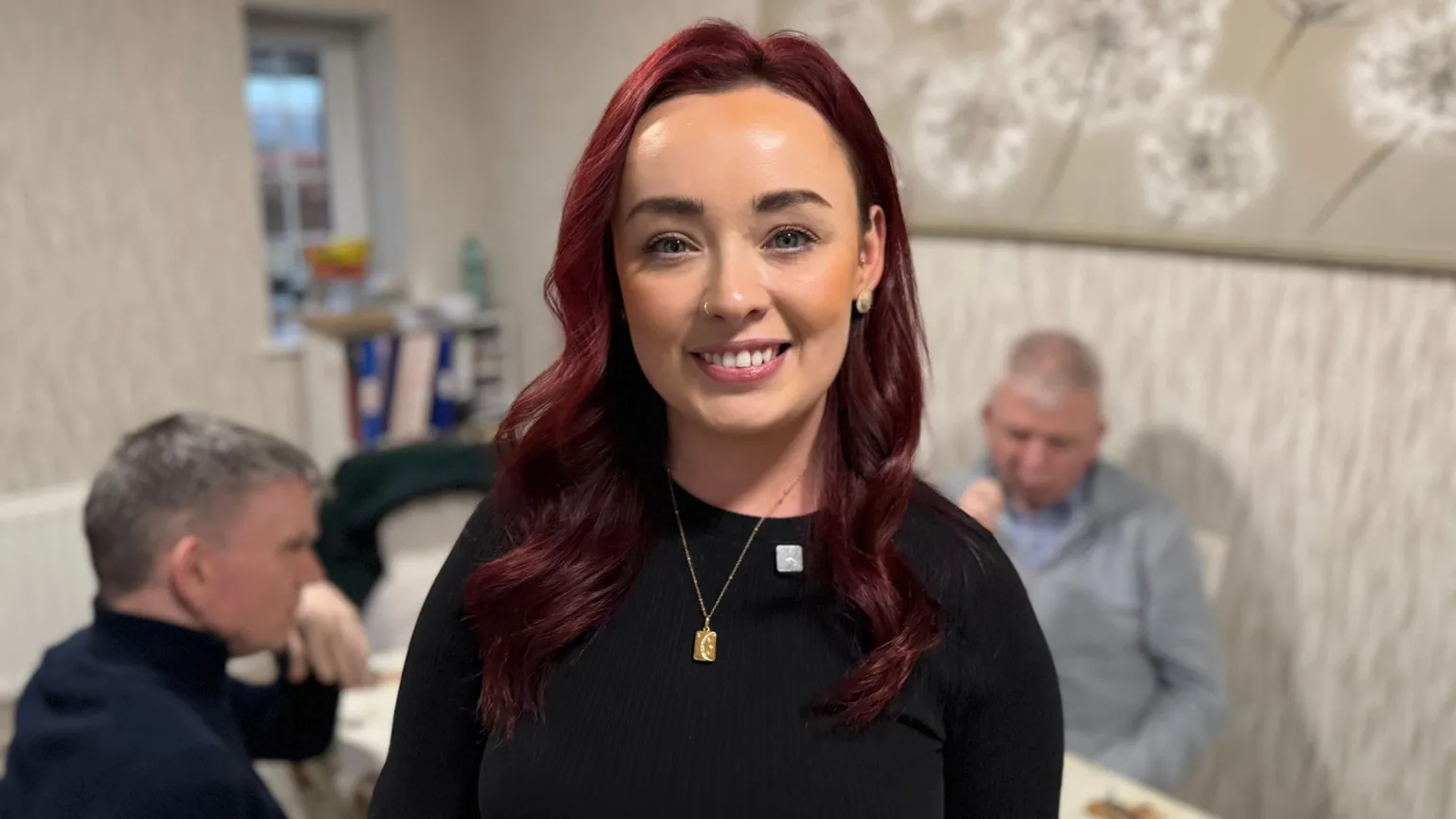 A young woman with red hair, which is curled and longer than shoulder length. She wears a black top and gold necklace. 