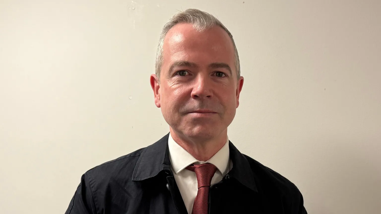 Pól Deeds has short grey hair swept back from his forehead and is wearing a dark coat, with a white shirt and red tie