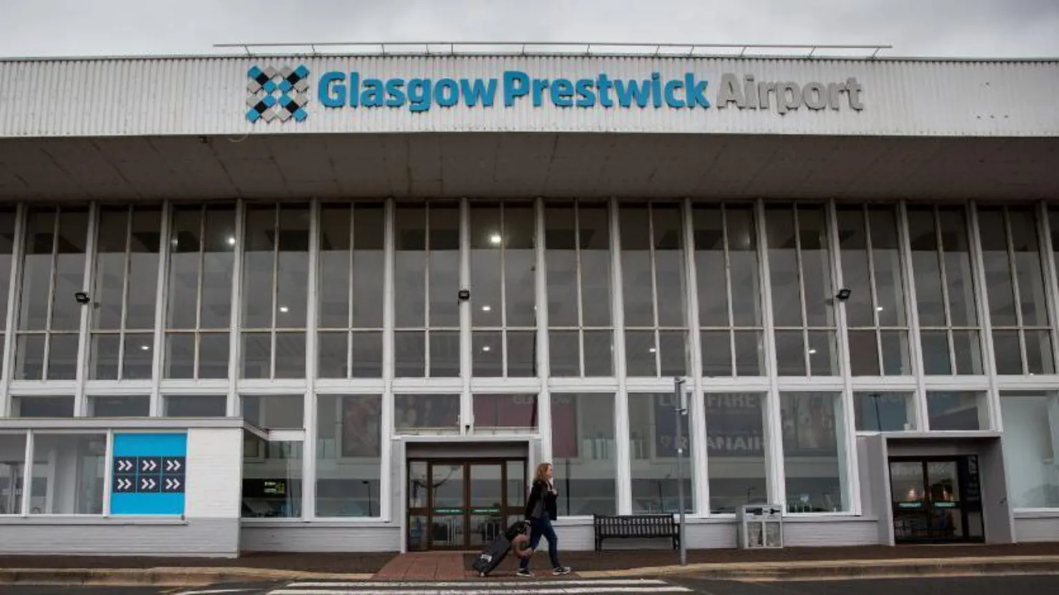  A view of the main entrance of Glasgow Prestwick Airport. There is a zebra crossing in the foreground, with a woman walking with a suitcase behind her on the other side of it. A large class building with a corrugated roof rises behind her. 