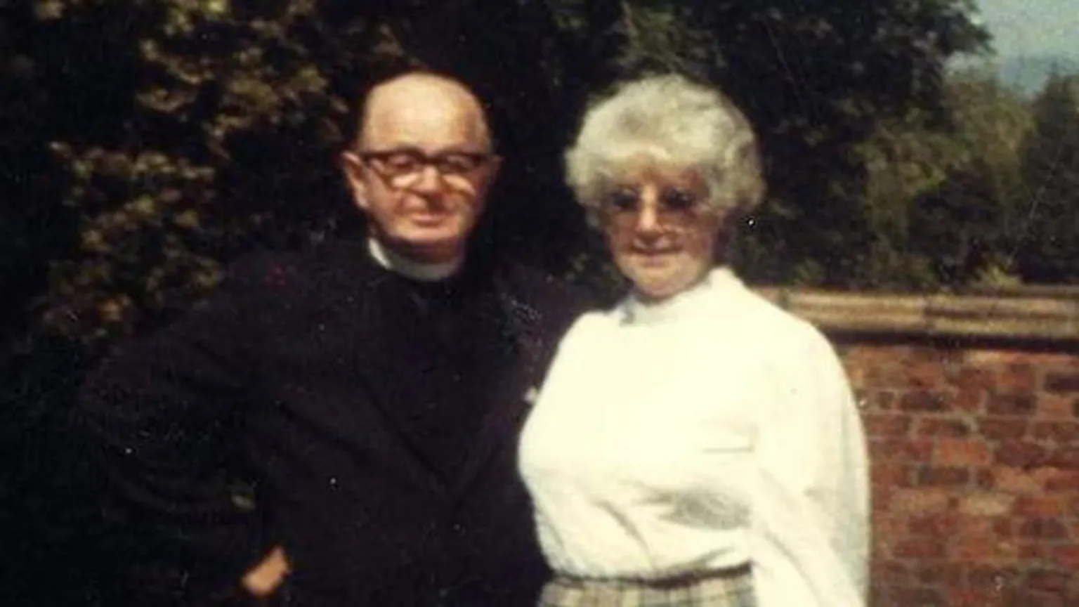 On the left, William Barrie is bald and is wearing glasses. He stands wearing a minister's uniform, with his arm around the back of Mary Barrie, who has grey hair and is wearing glasses and a white blouse. 