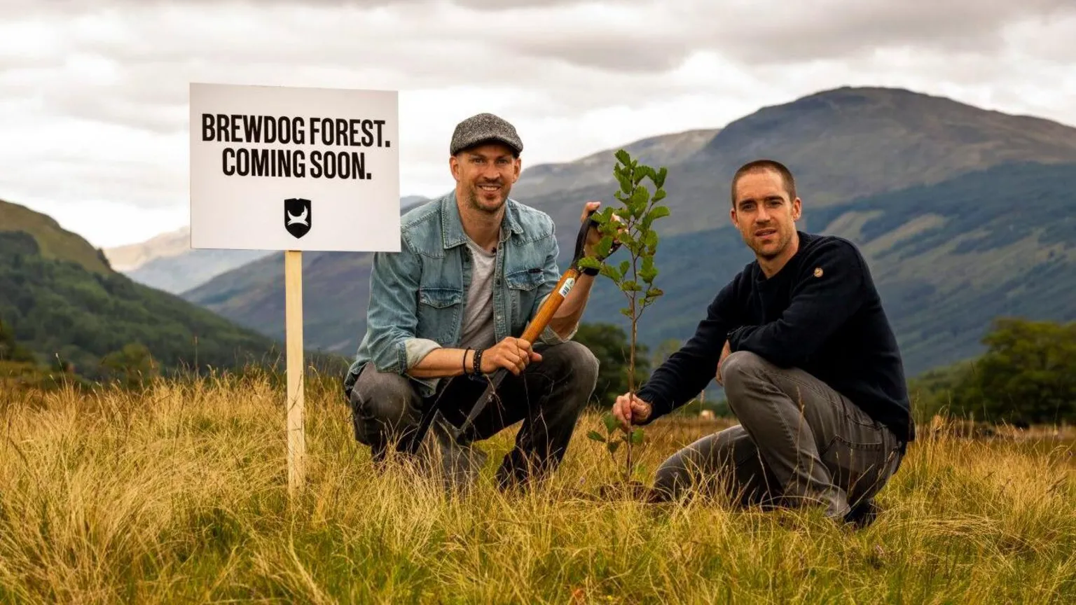 Brewdog James Watt in a cap and blue shirt holding a spade and Martin in a long sleeve navy top crouching down in a rural landscape with a sign beside them saying 'brewdog forst coming soon'