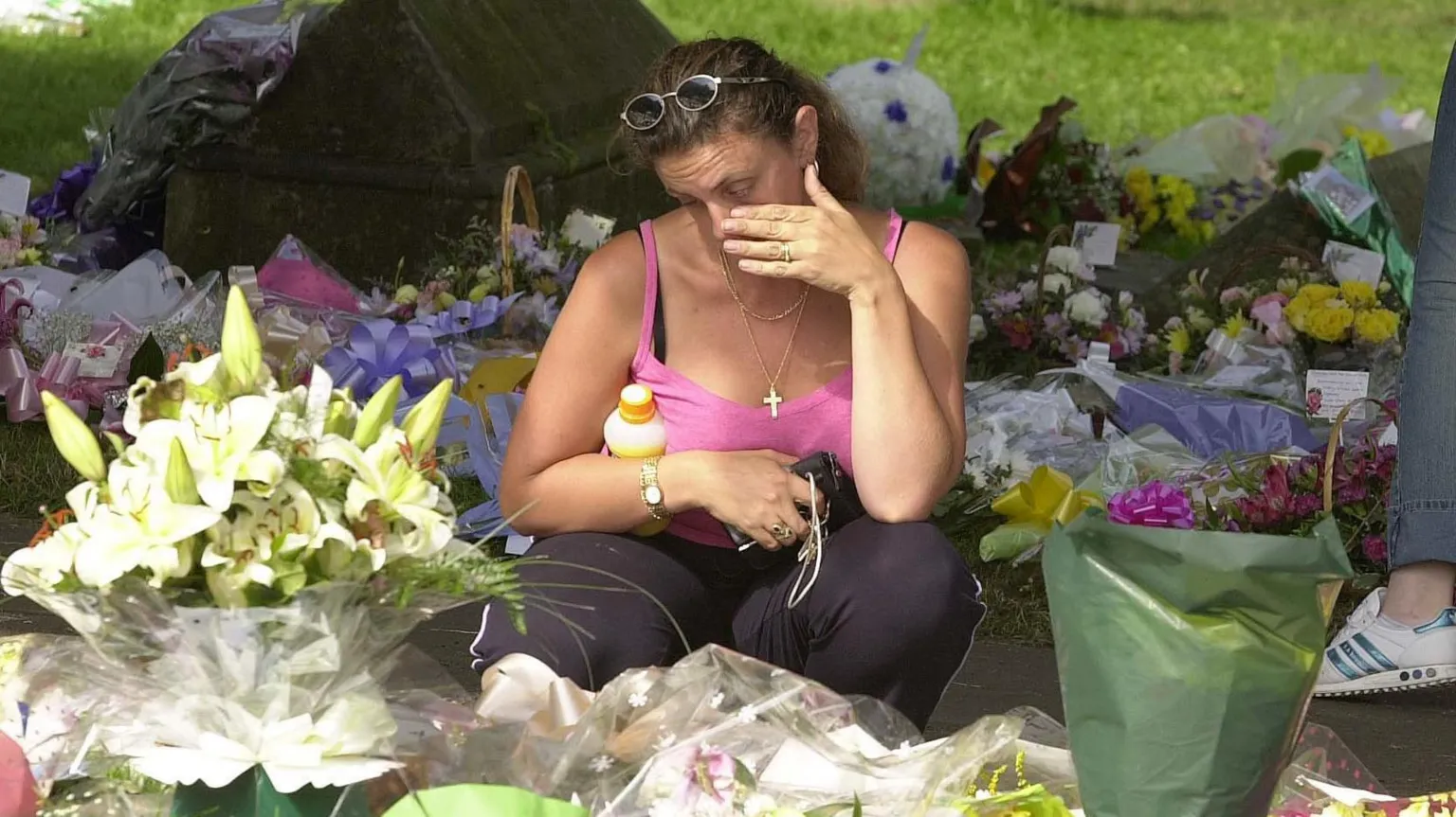  A woman wearing a pink vest top crouches down among lots of flowers laid in memory of Holly and Jessica. She is wiping a tear from her eye.