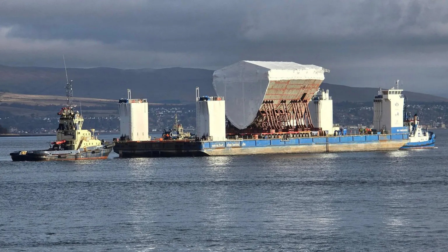 Christopher Brindle The front section of a ship, on a blue barge being pulled by tugs