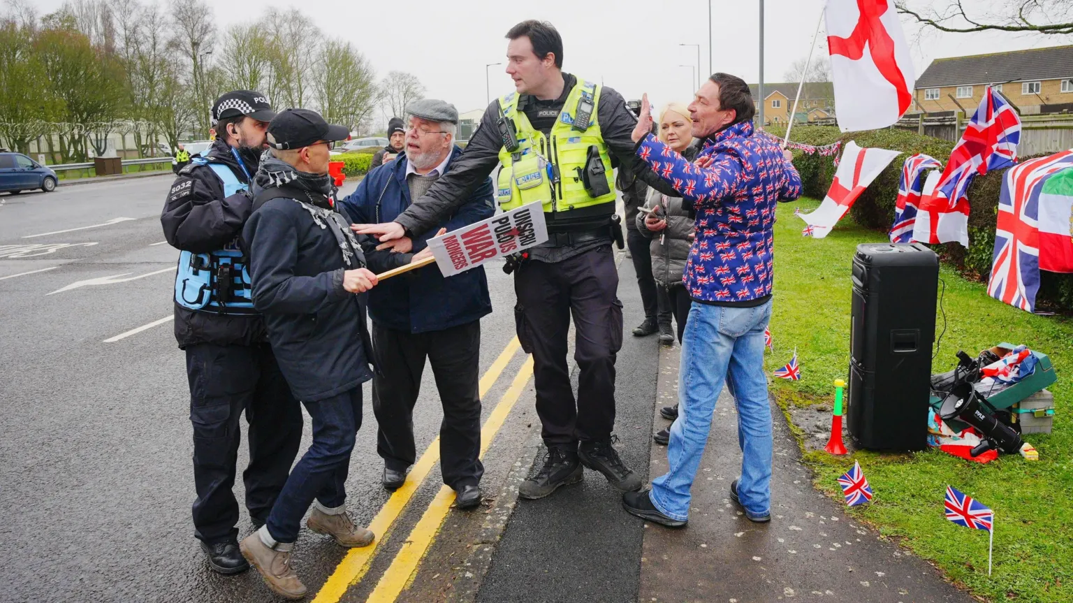  A police officer stands between an anti-war protester and a protester in support of the UK military. The anti-war protester holds a sign that reads 'unscrupulous war mongers' and the pro-UK military protester holds England's Saint George's flag.