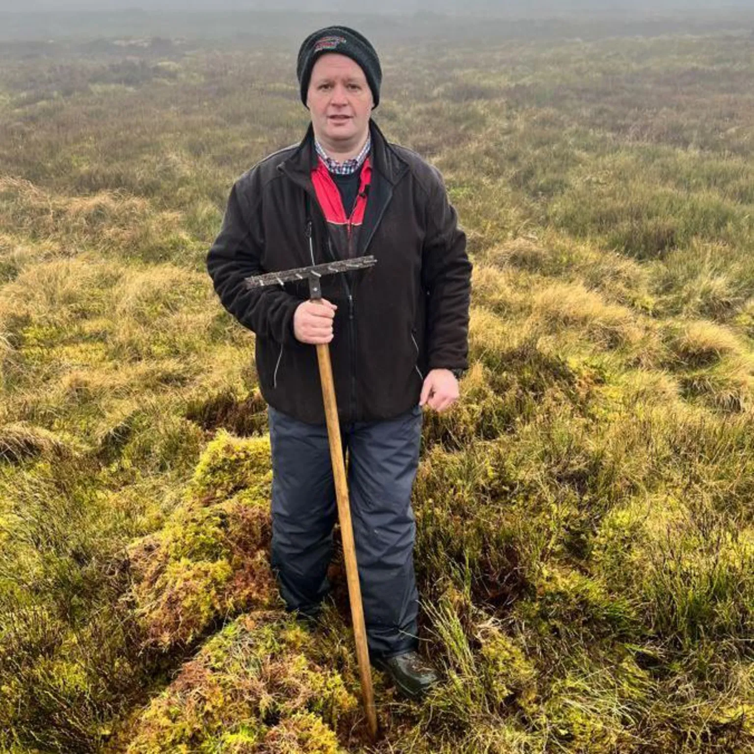 A man holding a rake stands in a field full of moss
