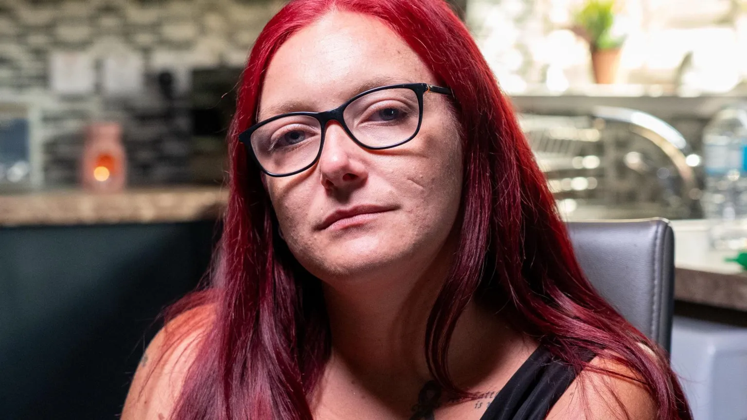 Yeti Television/BBC Cymru Wales Bethan Callaghan. She has dark reddish brown hair, blue eyes, and looks directly at the camera. She has black rimmed reading glasses and wears a black vest. She sits at the forefront of the camera and there is a blurred kitchen in the background. 