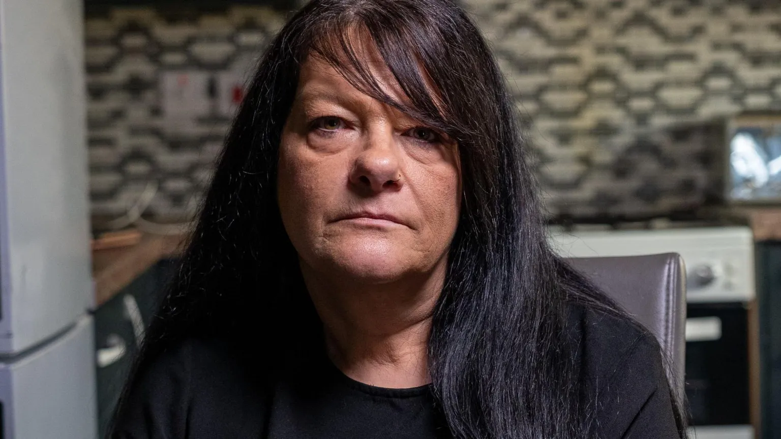 Yeti Television/BBC Cymru Wales Bethan Callaghan's mother, Gaynor. She has black long hair and a side fringe. She has brown eyes and stares blankly into the camera. She sits at the forefront of the photo and a blurred kitchen with black and white tiles can be seen in the background. 