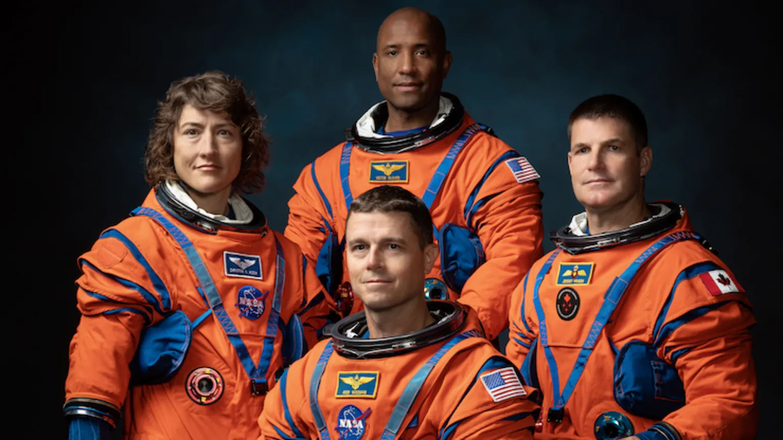 NASA The image shows four astronauts in bright orange space suits posing for a formal group portrait against a dark background. The suits are bulky with blue joints, straps and pockets, emphasising their technical design. Three astronauts stand behind one who is seated, all facing the camera. They all have fairly neutral expressions, keeping the focus on the uniforms. Mission-style patches and flags, including US and Canadian flags, are visible on their arms and chests. Soft, focused lighting makes the vivid orange suits stand out dramatically. 