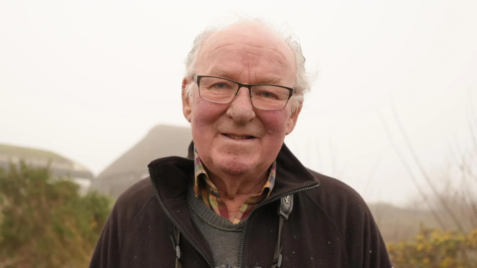 Shaun Whitmore/BBC Bernard Bishop, a man who is standing outside on the Cley Marshes reserve. The photograph has been taken on a foggy day with the background of the reserve difficult to make out clearly. He is wearing a grey jumper and chequered shirt with a black fleece over the top. He is looking directly at the camera and smiling.