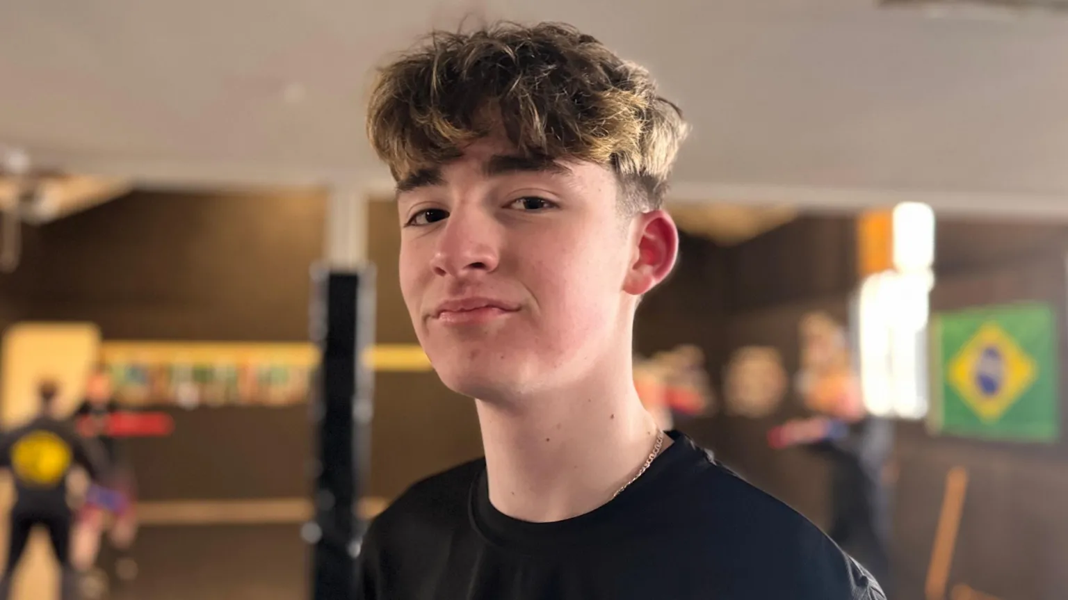 Loughlin is a young boy. He has medium-length brown hair with some blonde highlights. He is stood in the middle of a training room, but the background is blurred. He is turned slightly to the side and is smiling with his mouth closed at the camera. He is also wearing a black t-shirt.