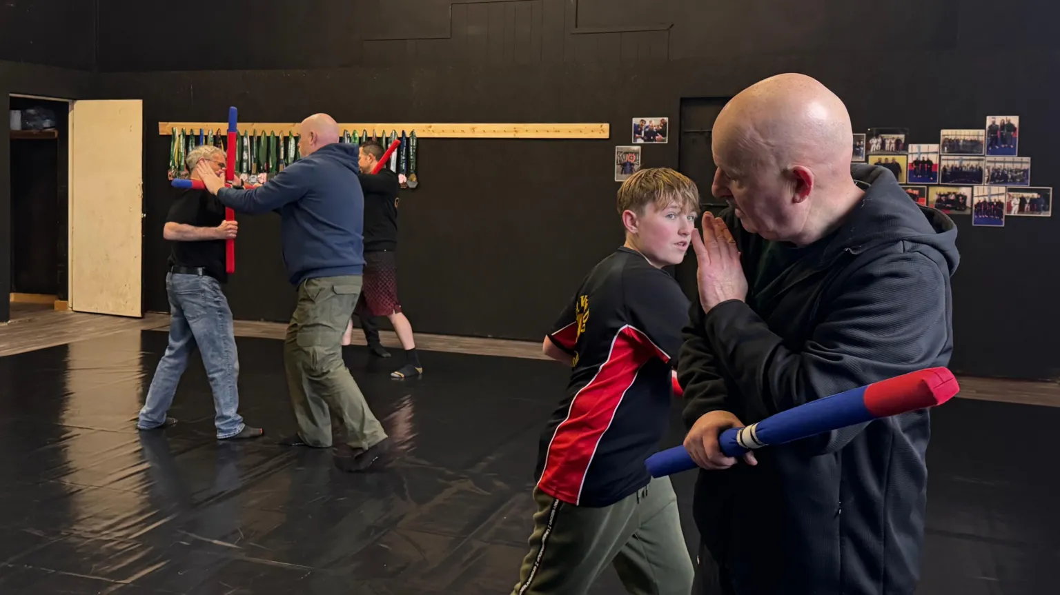 A number of different pairings engage in a practice bataireacht fight. There are a couple of older men, and a young boy. They are stood in the middle of a martial arts studio, which has black padded mats and walls. They are holding red and blue foam sticks. 