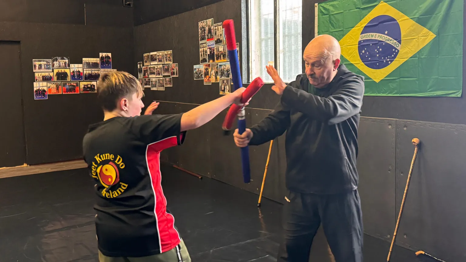 A young boy battles with Martin O'Neill, an older bald man who is wearing a black tracksuit set. The pair are using foam sticks to battle each other. They are stood on black mats in the middle of a martial arts studio, which has a Brazilian flag on one wall, and a number of photographs on another.
