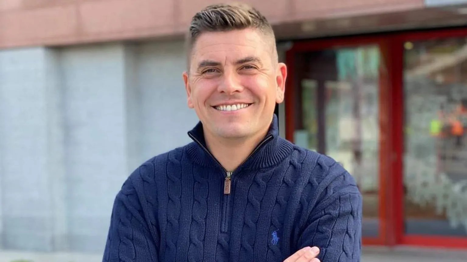 Sinn Féin Danny Baker. He has light brown hair, wearing a navy half-zip, with his arms folded. He is smiling at the camera.