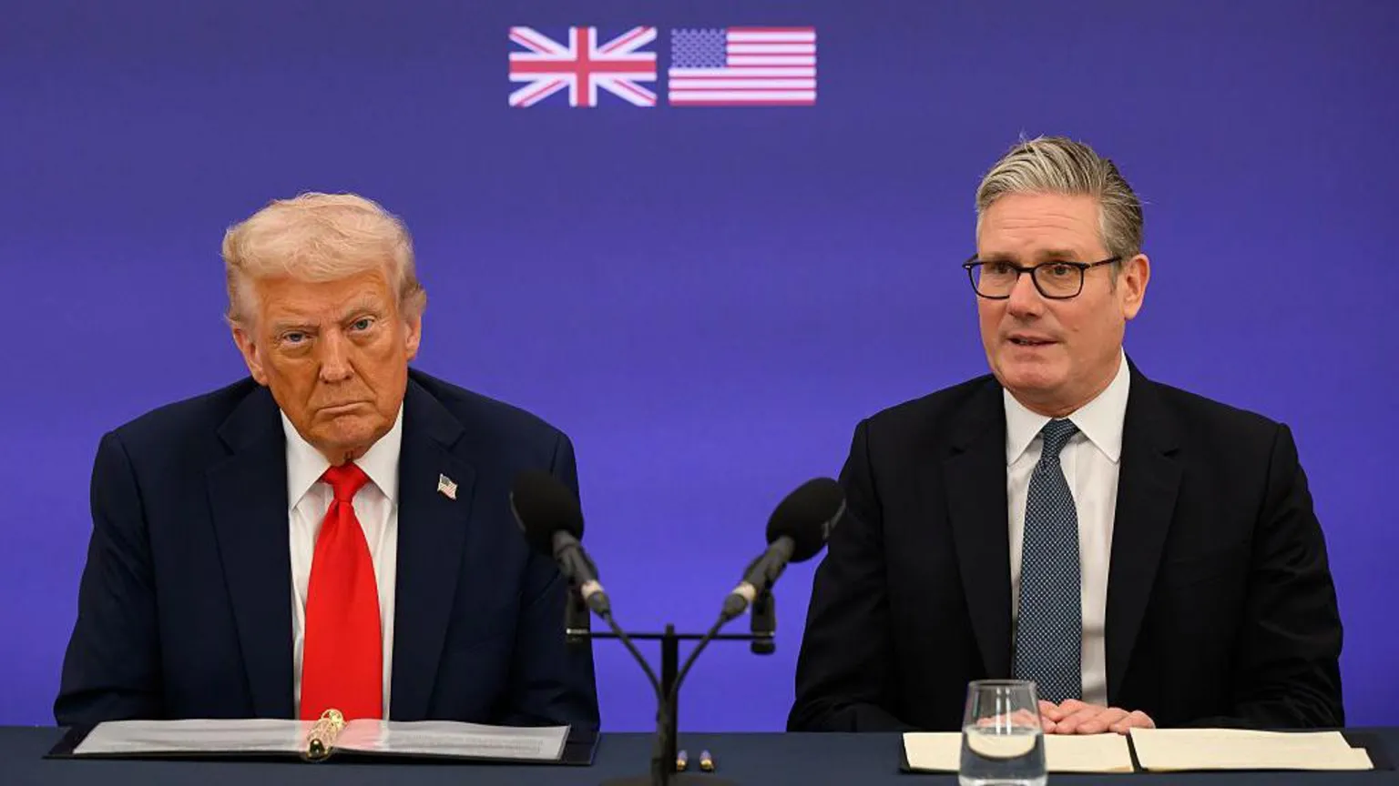  Trump and Starmer sit at black desk with black microphones. A purple coloured screen behind them has small UK and US flags pressed into it. Starmer is wearing a black suit and patterned tie with a white shirt. Starmer is looking ahead with his hands crossed over one another. Trump is looking straight ahead and appears to be scowling. He is wearing a navy suit with a red tie and a white shirt.