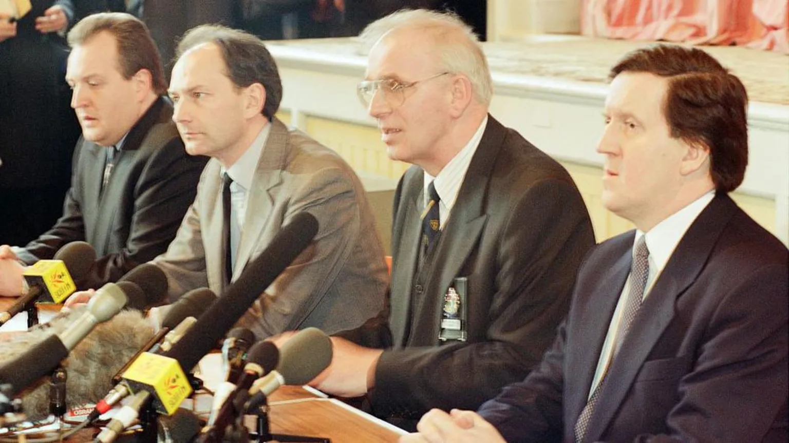 Mirrorpix via Four men in dark suits and ties sit in a row at a table. They look very shocked and serious. There are microphones on the table.