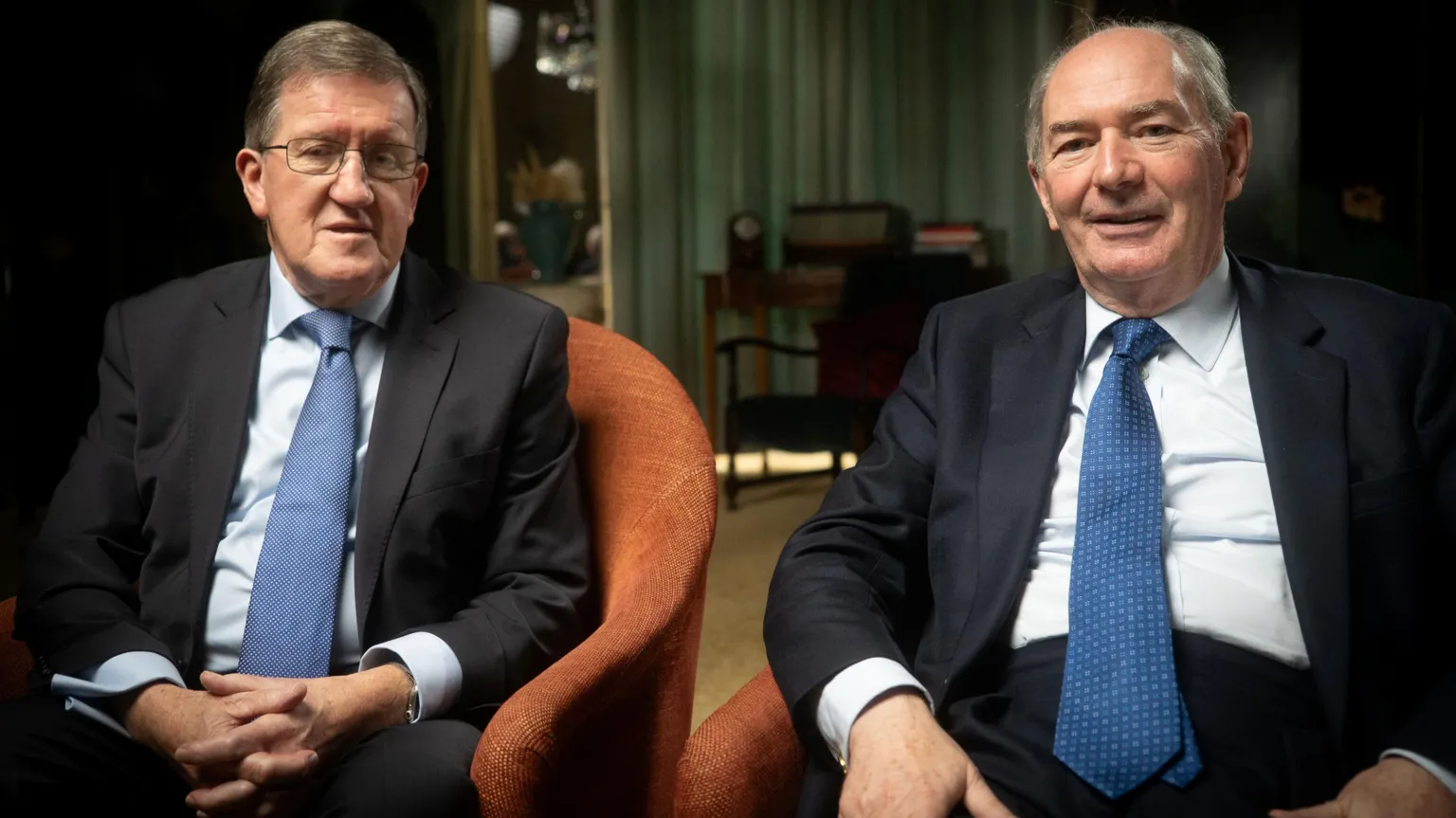 IWC Media George Robertson and Michael Forsyth sit side-by-side in a room with armchairs and draped green curtains. They are in similar dark suits, light blue shirts and mid-blue silk ties.