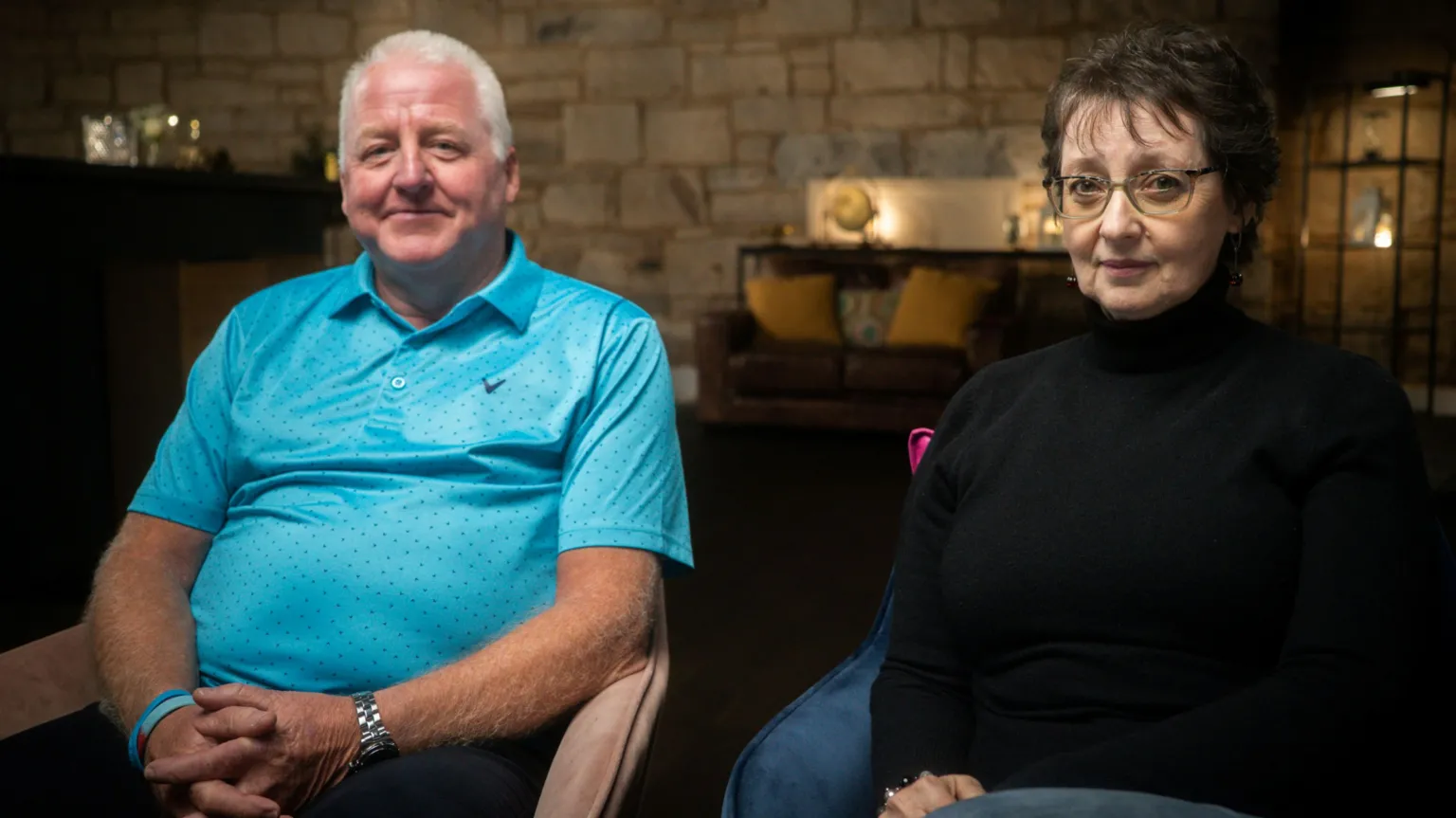 IWC Media Kenny Ross in a vivid blue golf-styled shirt and Pam Ross in a black polo-neck jumper and jeans, sit on chairs in a stone-walled room. They are looking at the camera and Kenny is smiling slightly. Pam has glasses.