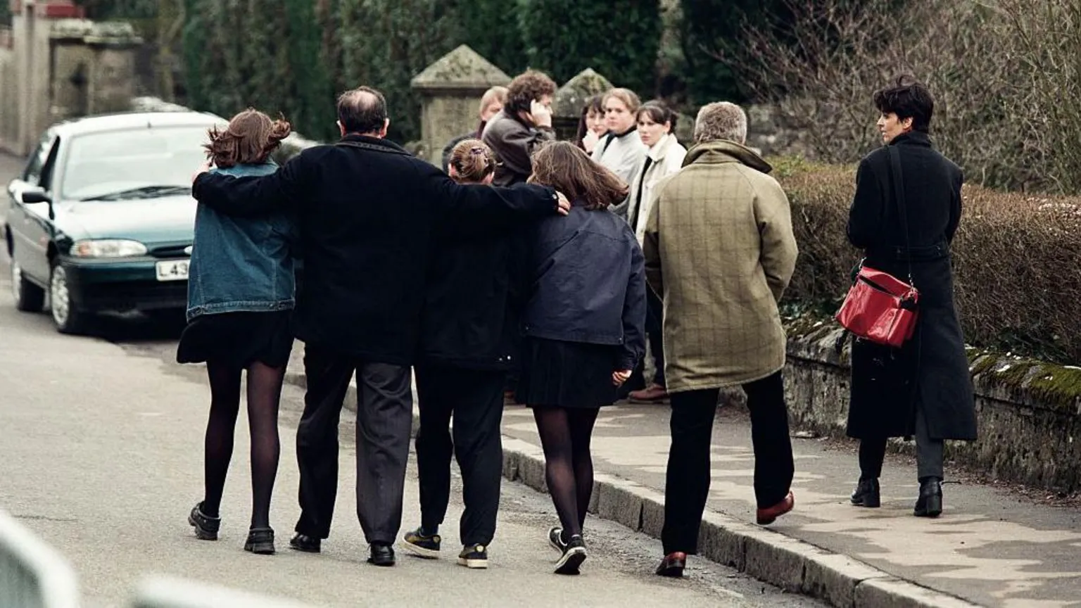  A group of three teenage girls walk away from us down a stone-walled road. A man between them has his arms protectively draped over their shoulders. Others stand in groups beyond them. A man and a woman are also beside them, walking away.
