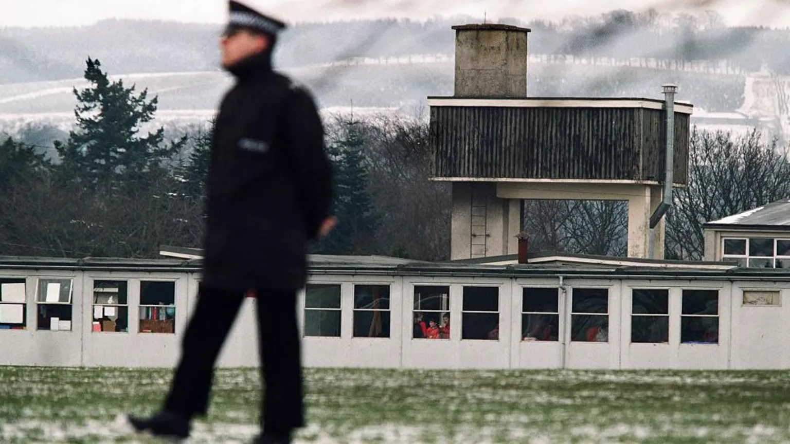  A police man in full winter coat and a peaked cap walks in front of brutalist concrete and wood buildings. There is snow on the ground and a lot of snow on hills in the background.