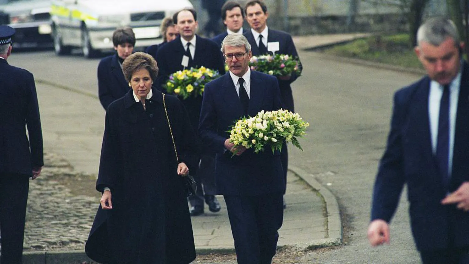  A group of seven people walk towards camera down a street with police cars in the background. John Major, Michael Forsyth and Tony Blair can be seen carrying wreathes. George Robertson can be seen in the background. All are in dark suits, white shirts and black ties. 
