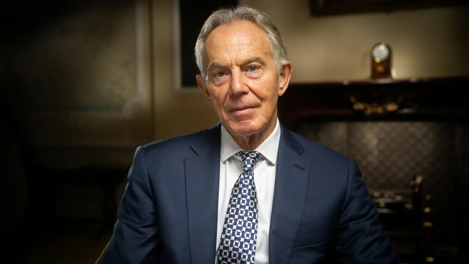 IWC Media Sir Tony Blair, in blue suit, white shirt and blue and white checked tie, sitting in front of a clock on a mantlepiece in an atmospherically-lit room.