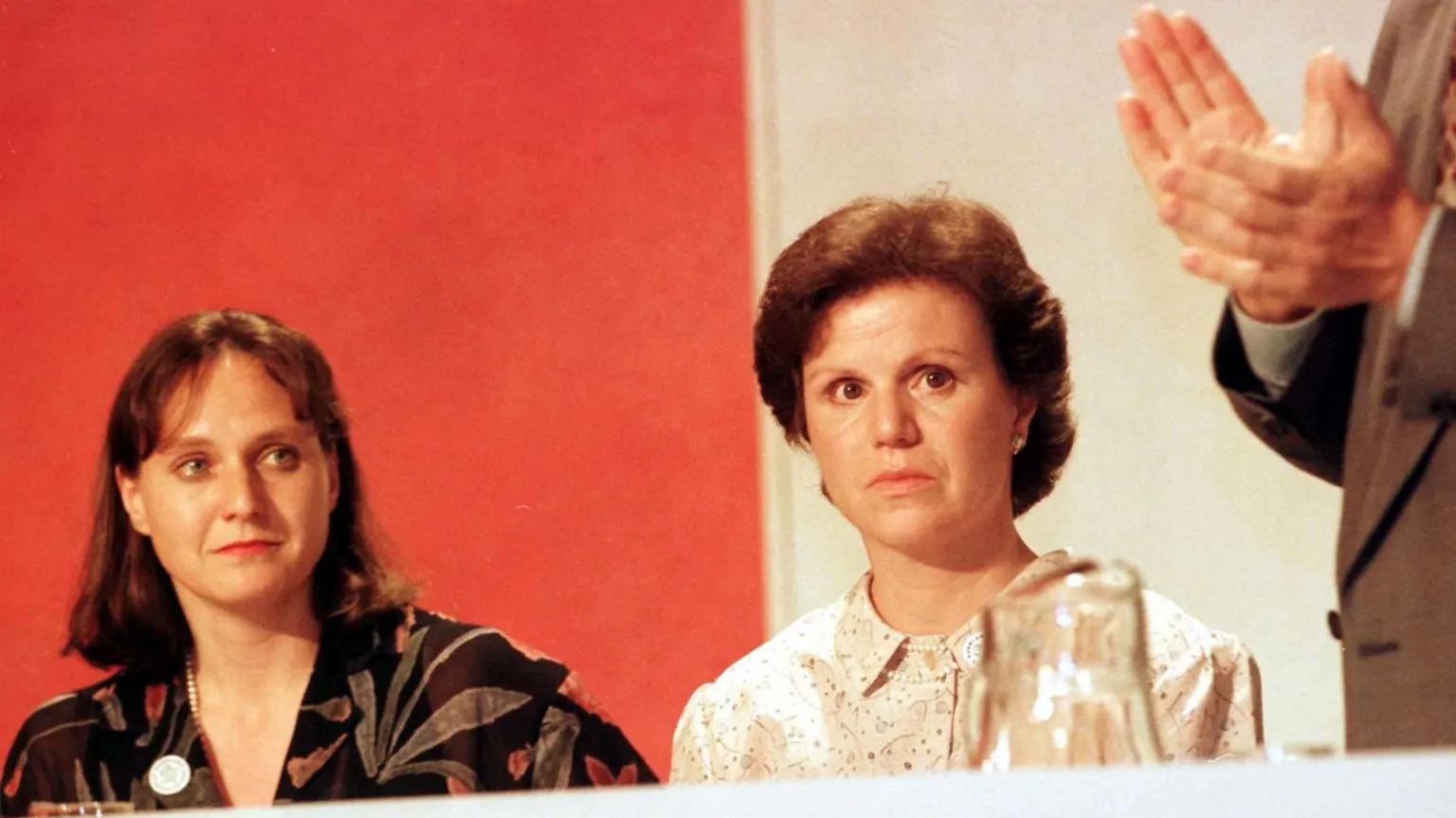  Rosemary Hunter in patterned brown top and Ann Pearston in a cream top are watching a man in a beige suit on edge of frame talk. They are listening intently. There is a red and white backdrop and some glasses in the extreme foreground.