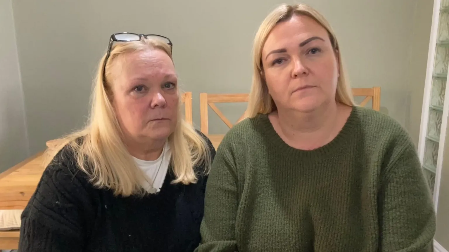 Two women look at the camera. The one on the left has glasses on top of her head and blonde, shoulder-length hair. She is wearing a black jumper with a white t-shirt underneath. The one on the right has a green jumper on and long blonde hair. Their expression is neutral. There is an oak table and chairs behind them. 