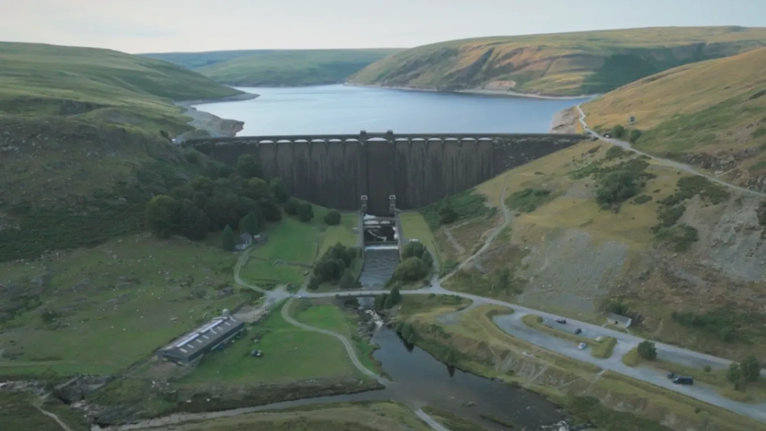 BBC Crimewatch A drone photo taken downstream from the Claerwen Reservoir, showing the dam with the reservoir behind it. In the foreground is a river, surrounded by green fields and a barn. Behind that is a large stone dam with arches along the top. Beyond that is a large blue reservoir, surrounded by bare green upland hills.