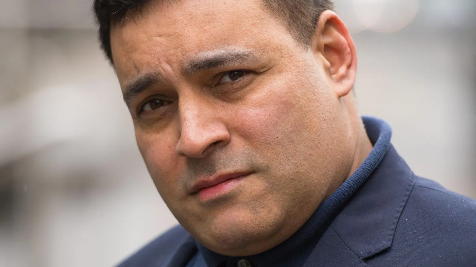  A close up picture of a man with black hair wearing a navy top. The background is out of focus.